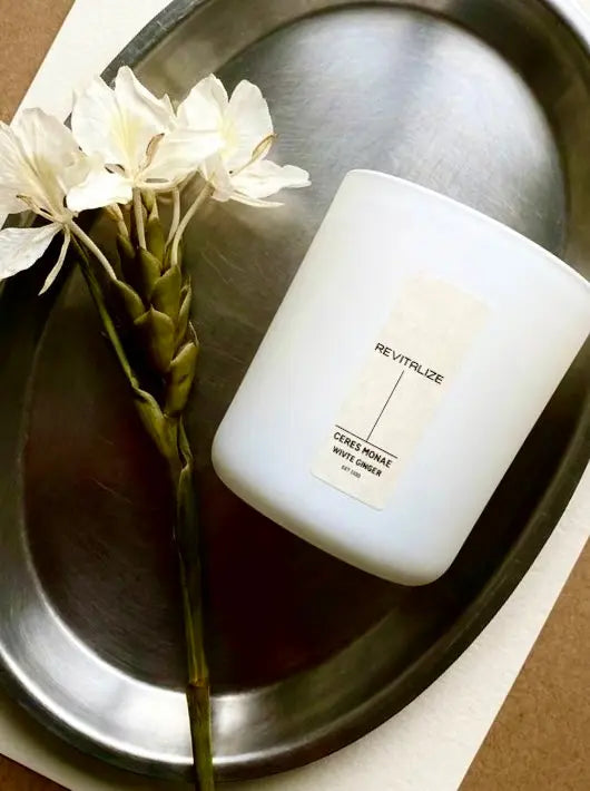 White candle labeled 'Revitalize' with white flowers on a metallic surface